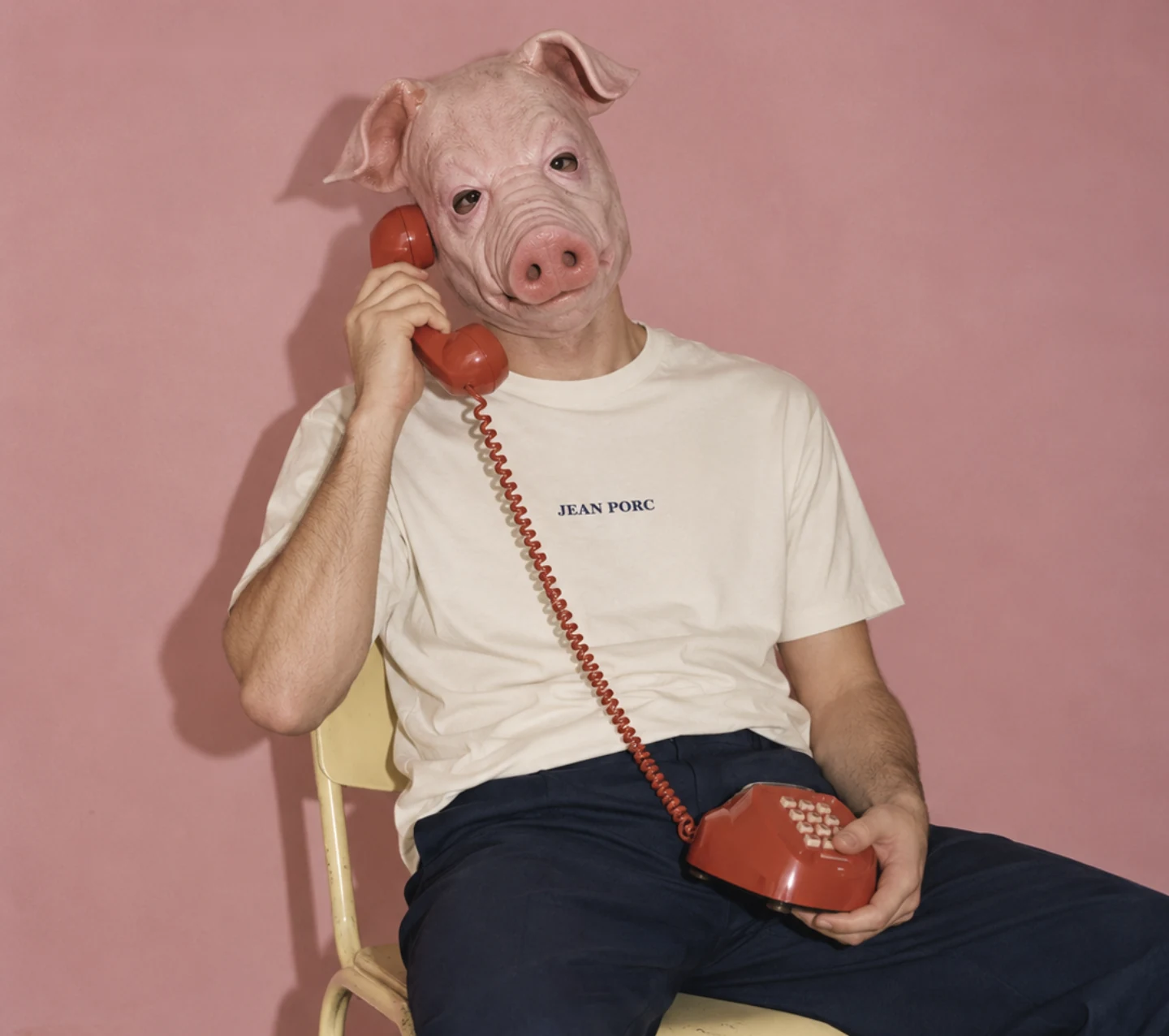 A man in a pig mask wearing a crème Jean Porc tee, talking on a red rotary phone against a pink wall